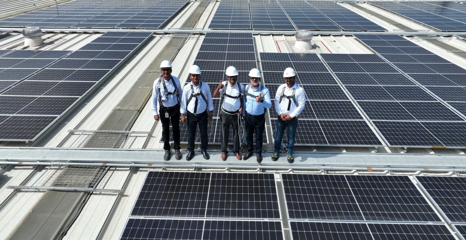 Four individuals wearing white hard hats and safety harnesses stand on a rooftop covered with solar panels. They are smiling and posing for the photo, with solar panels stretching out in rows behind them, indicating a large solar installation.
