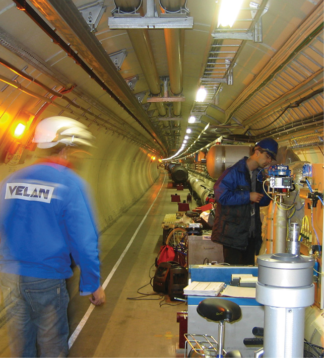 An engineer in protective gear works on machinery inside a long, well-lit tunnel with pipes running along the walls and ceiling. Another person, blurry in motion, wears a jacket with "VELAN" on the back and a white hard hat while walking away from the machinery.