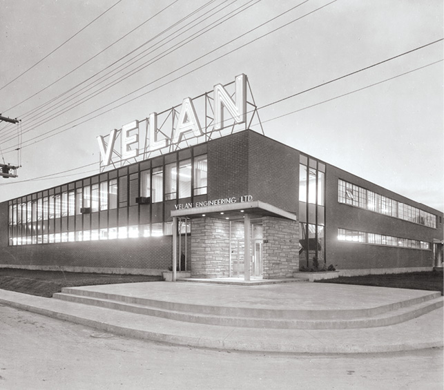 Black and white photo of a large industrial building with "VELAN" in large glowing letters on the roof. "Velan Engineering Ltd." is visible above the entrance. The building has numerous windows, with a set of stairs leading up to the front door. Power lines run overhead.