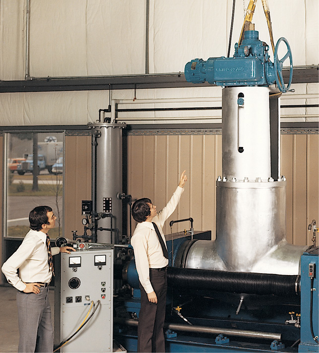 Two men in dress shirts and ties stand next to a large industrial valve system. One man is pointing towards the top of the valve, which is connected to a blue actuator or motor and suspended by ropes. The setting appears to be an industrial facility with machinery.