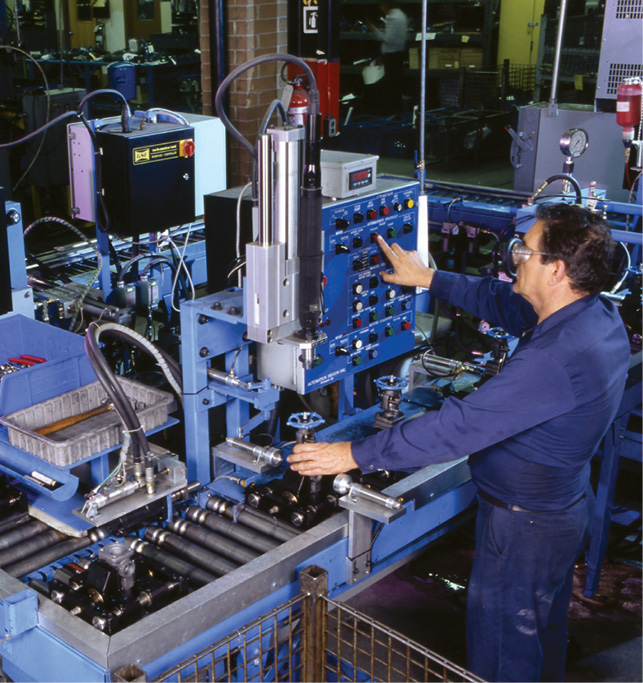 A worker in blue overalls operates a complex industrial machine with various buttons, knobs, and displays. The machine appears to be part of a larger assembly line in a factory setting. The worker is adjusting settings on the control panel.