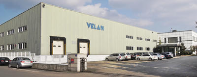 Large industrial building with "VELAN" on the facade. There are multiple loading docks and a parking lot filled with cars. The sky is partly cloudy.