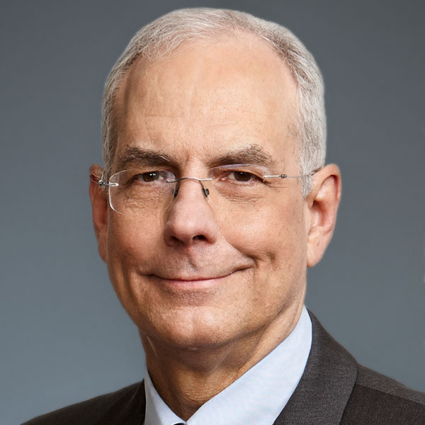 A person with short gray hair and glasses is wearing a dark suit and light shirt, standing against a plain, gray background.