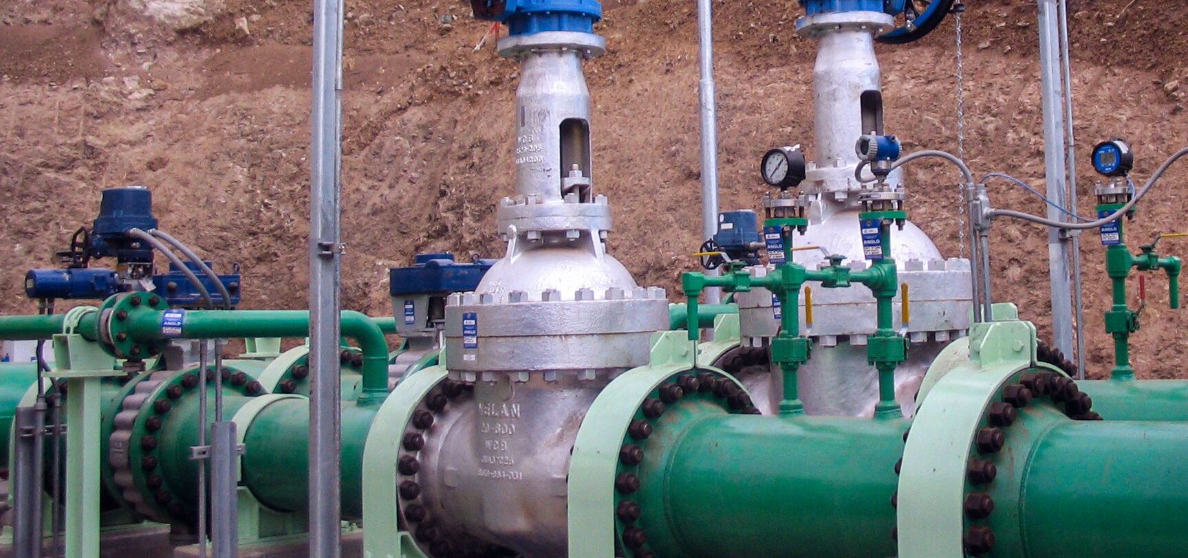 Several large metal industrial valves and pipelines, painted green, are installed outdoors against an earthen background. Pressure gauges, blue actuators, and green-handled controls are attached to the system.