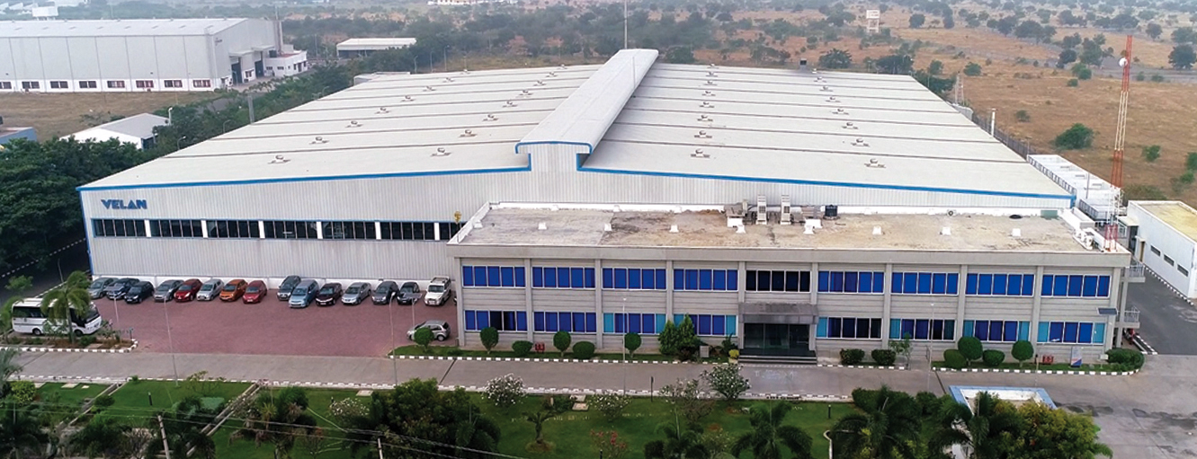 A large industrial building with "VELAN" on the side, featuring a spacious parking area filled with cars. The structure has a long, white roof and numerous blue windows. Trees and greenery surround the building under a clear sky.