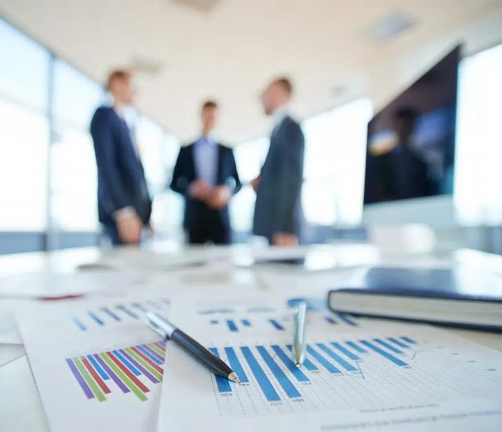 Three business professionals stand discussing in a bright office space. Out-of-focus charts and pens are visible on a table in the foreground, emphasizing a meeting or presentation setting.