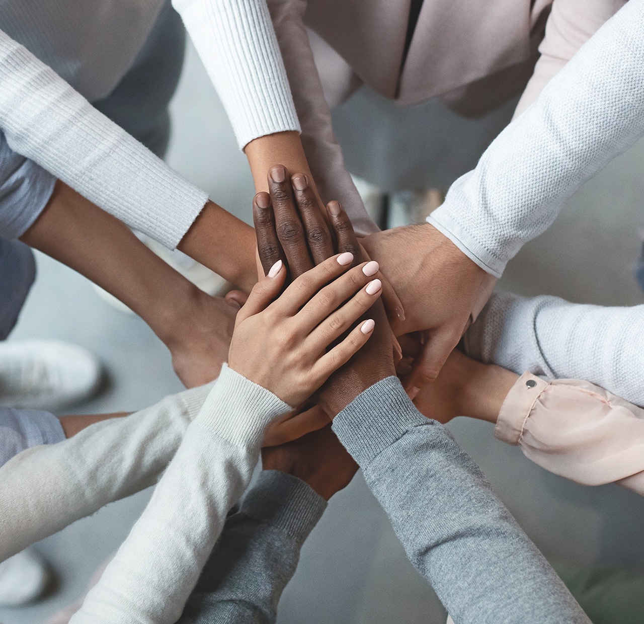 A diverse group of eight people stand in a circle, extending their arms inward to place their hands on top of each other, symbolizing unity and teamwork. They are wearing casual, long-sleeved clothing in neutral tones.