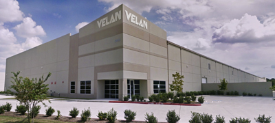 Exterior view of a large industrial building with "Velan" written on the facade. The building has a modern design, surrounded by a paved area and small landscaped bushes under a partly cloudy sky.