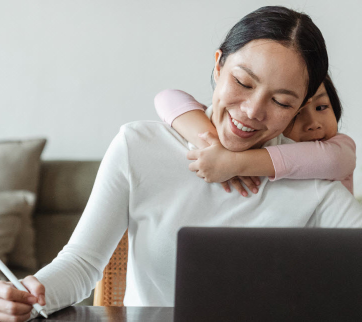A woman smiles while writing with a stylus at a desk with a laptop in front of her. She is being hugged from behind by a child, who also smiles. The background features a sofa, and the setting appears to be a home.