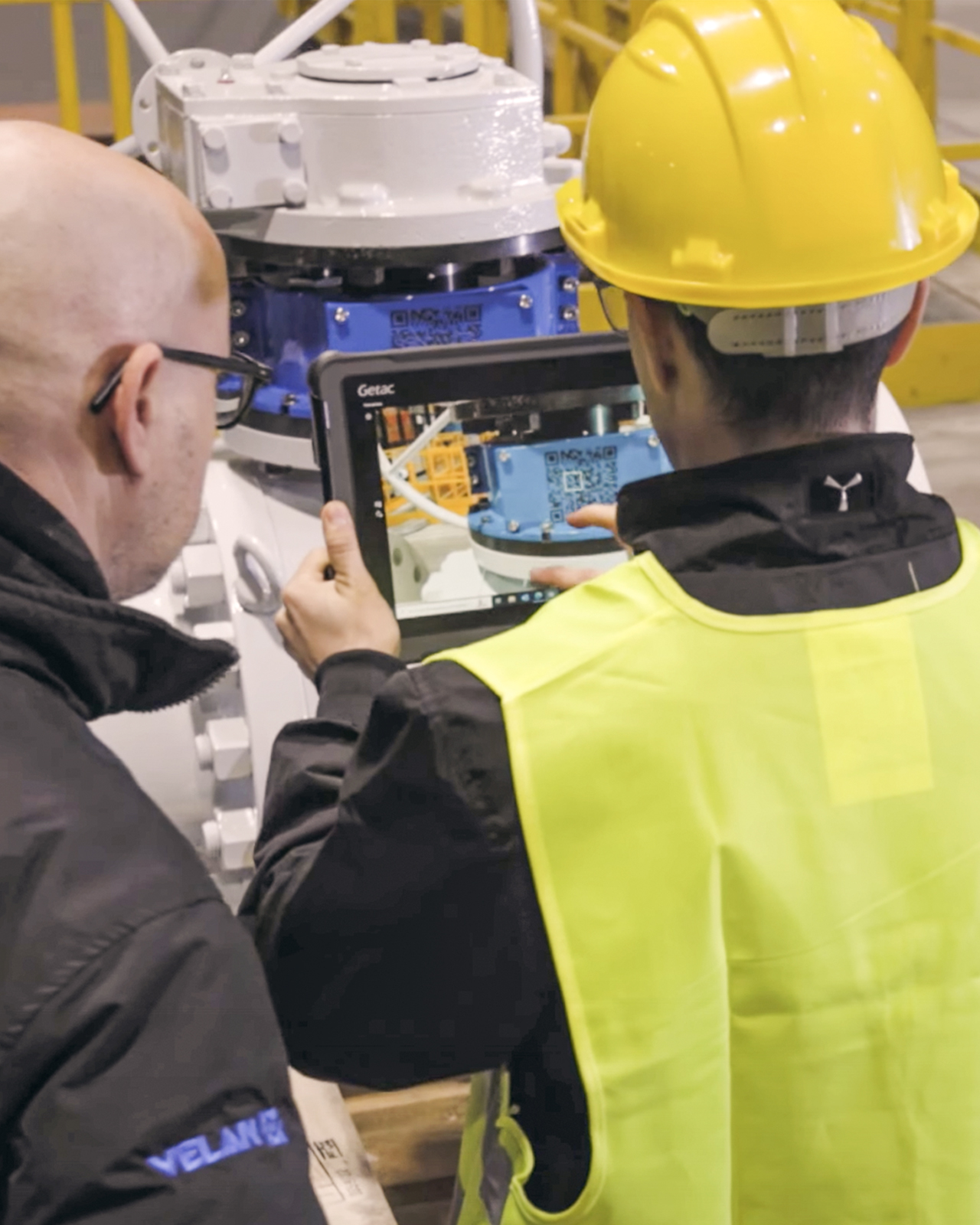 Two engineers, wearing protective helmets and high-visibility vests, inspect machinery using a tablet. One engineer holds the tablet, displaying technical details, while the other looks on. They are in an industrial setting with equipment and structures in the background.