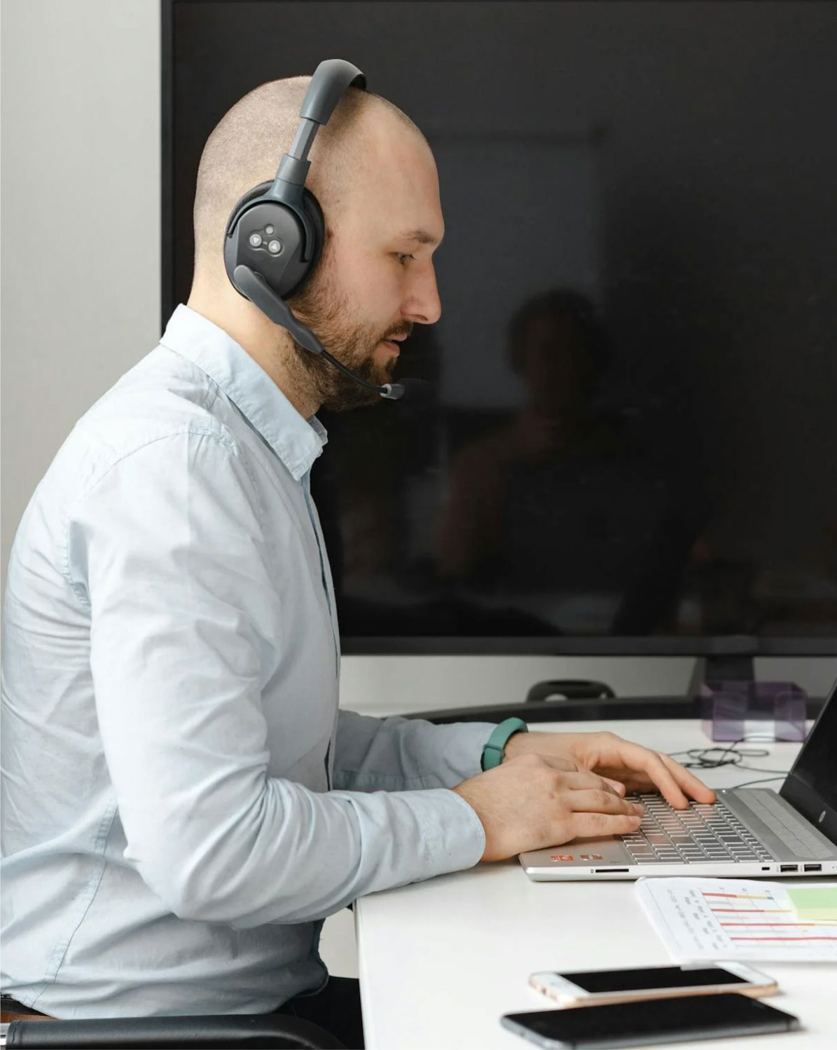 A man wearing a headset works at a laptop in an office setting. A smartphone is on the desk beside a notebook and a document. A large screen is in the background.