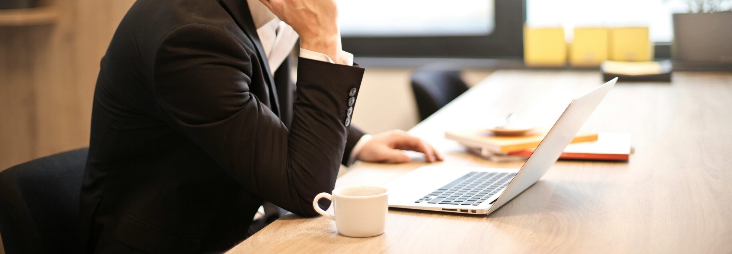 A man in a suit is sitting at a desk, talking on a smartphone while looking at a laptop. There is a white coffee cup on the desk, and the background shows a large window with natural light coming in. The workspace appears professional and minimalistic.