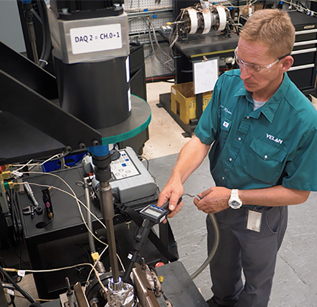 A man wearing safety glasses and a green shirt operates machinery in a workshop. He is using a handheld device to take measurements from the machine. The room is filled with various industrial equipment and tools in the background.